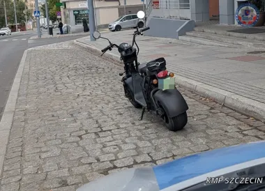 Czarny elektryczny skuter typu chopper zaparkowany na brukowanej nawierzchni przy chodniku w centrum miasta, w dolnym rogu widać fragment policyjnego radiowozu.